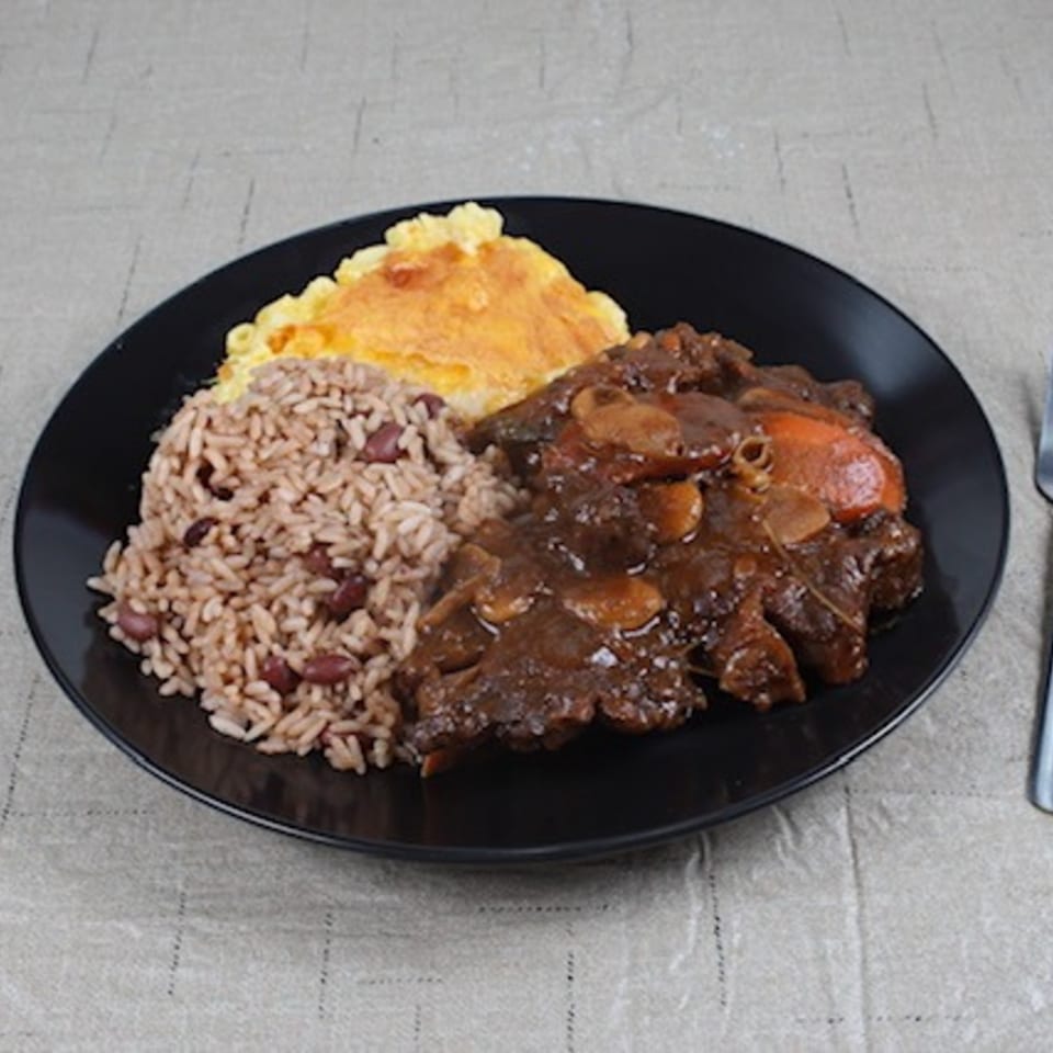Island Flavors' Jamaican oxtail stew, rice and peas, and baked macaroni and cheese on a black plate.
