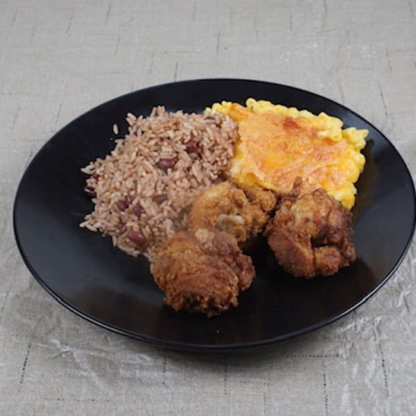 A black plate of Jamaican food with rice and peas, fried chicken, and baked macaroni and cheese.